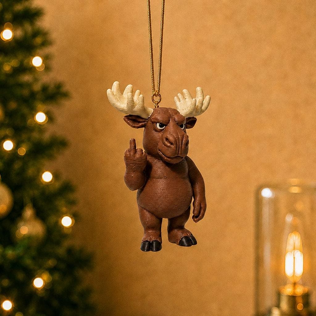 Funny brown moose Christmas ornament hanging from a pine branch beside a decorated tree and warm lamp, with cream antlers and a raised middle finger.