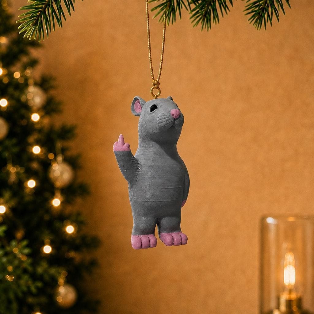 Funny grey rat ornament hanging beside a decorated Christmas tree and warm lamp, with pink ears, feet and a raised middle finger.