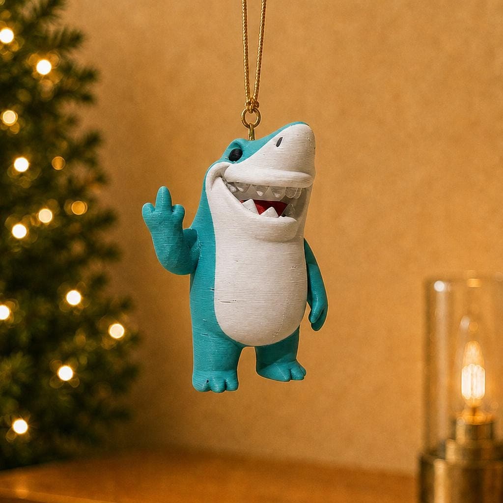 Funny blue shark Christmas ornament hanging beside a decorated tree and warm lamp, with a white belly, toothy grin and raised middle finger.