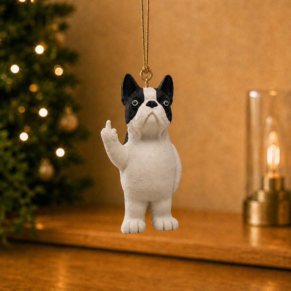Funny Boston Terrier ornament hanging beside a decorated Christmas tree and warm lamp, with black and white markings and a raised middle finger.
