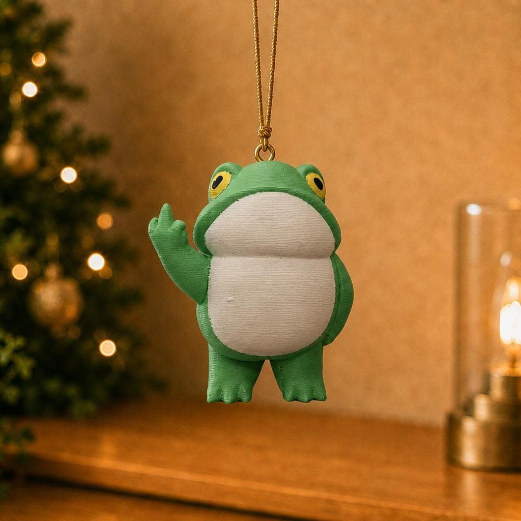 Funny green frog ornament hanging beside a decorated Christmas tree and warm lamp, with a round white belly and raised middle finger.