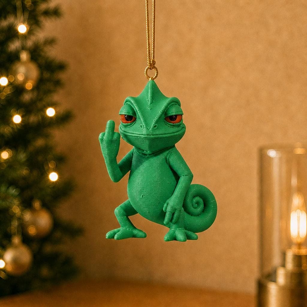 Funny green chameleon ornament hanging beside a decorated Christmas tree and warm lamp, with red eyes, a curled tail and raised middle finger.