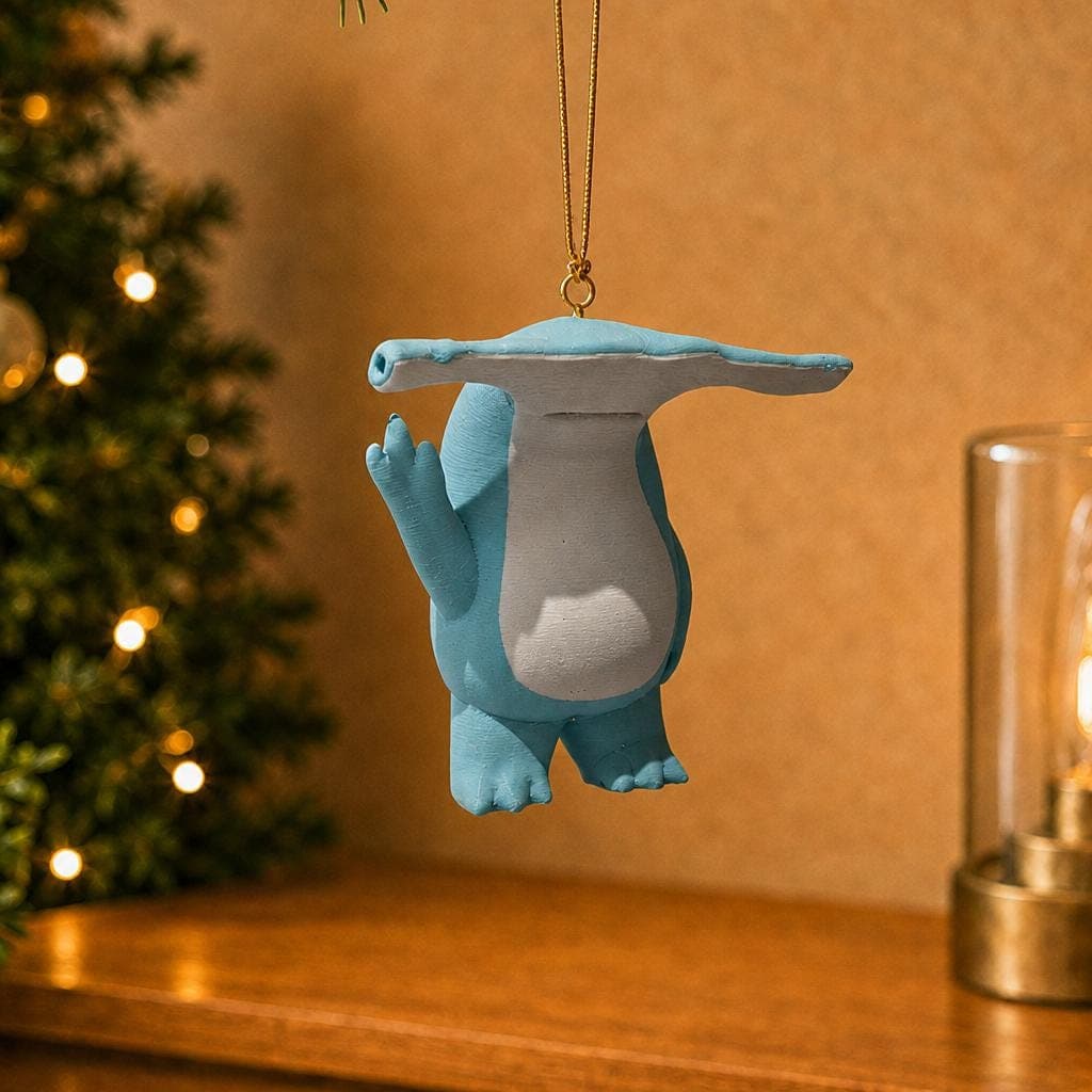 Funny blue hammerhead shark ornament hanging beside a decorated Christmas tree and warm lamp, with a white belly and raised middle finger.